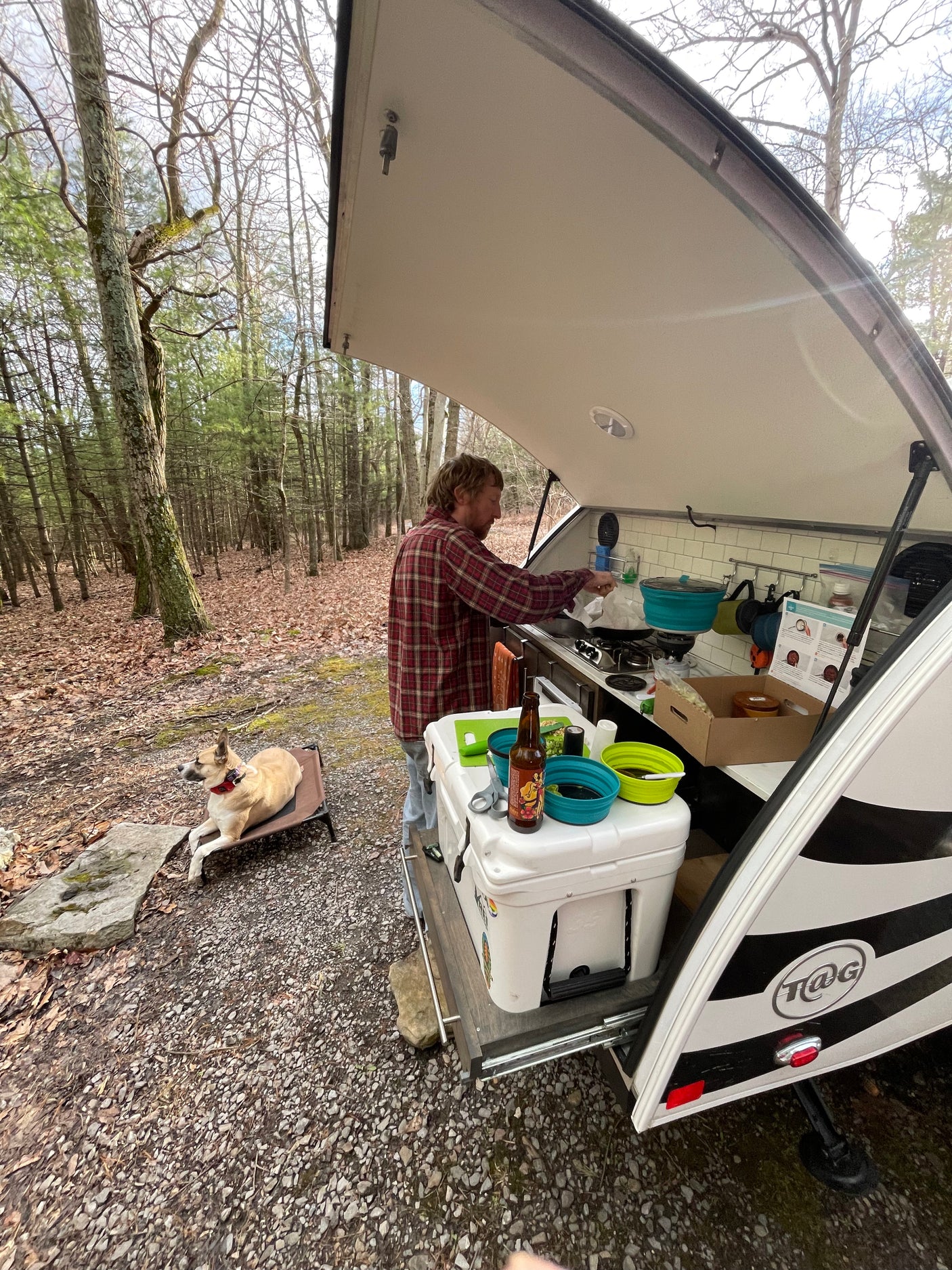 RV rental camping: A person cooks at their teardrop trailer's open kitchen while a dog lounges on a bed.