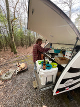RV rental camping: A person cooks at their teardrop trailer's open kitchen while a dog lounges on a bed.