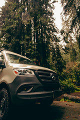 Experience nature with this RV rental. A modern van in a wet, dense forest, rain streaking the tall trees.