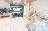 Inside a bright RV rental: features a bed, kitchen, overhead storage, and rotating front seats forming a dining area.