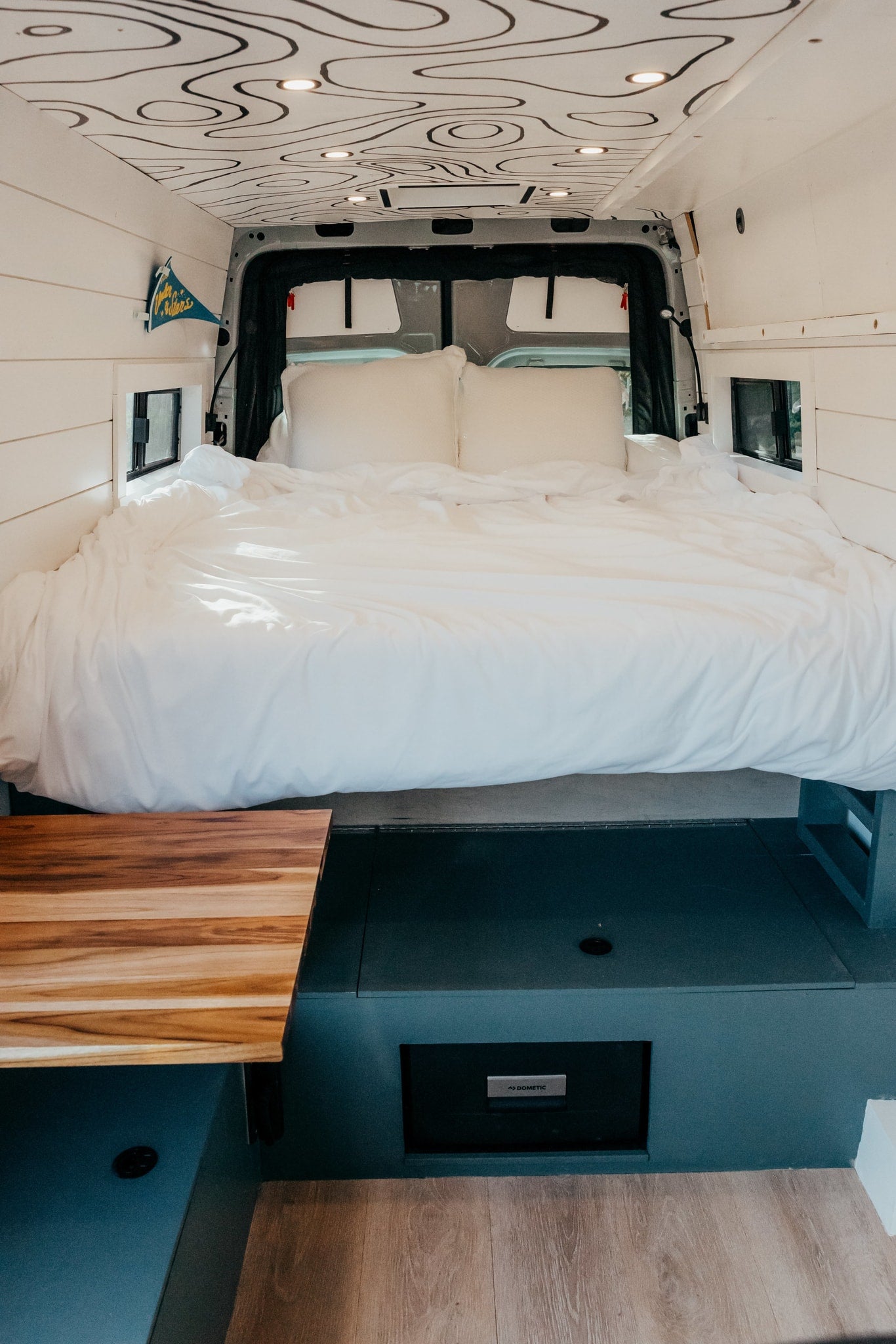  with a wooden table & upholstered bench in the foreground. Topographic ceiling.|Look up in this RV rental: a modern camper van interior with a ceiling fan — photo 7