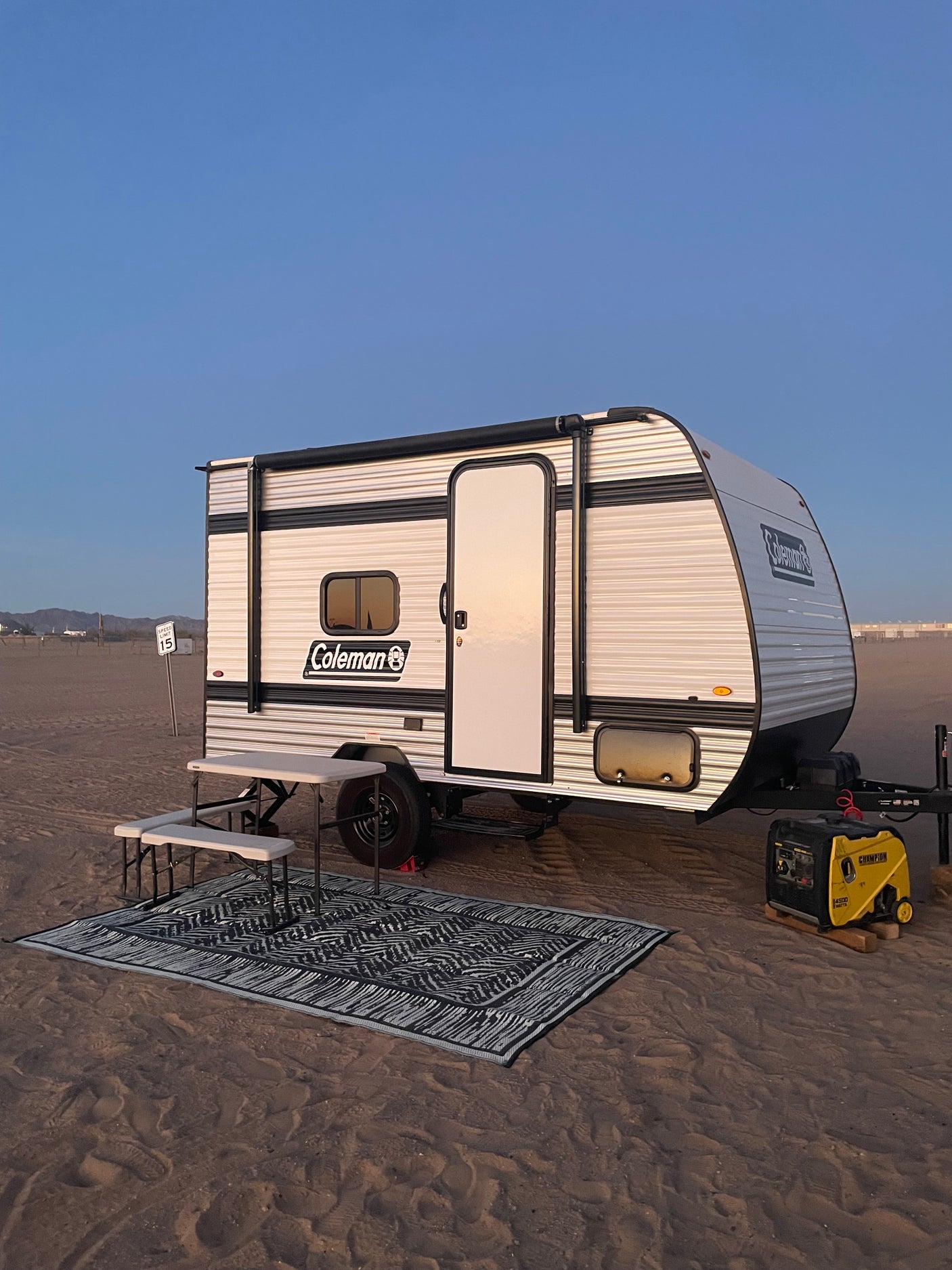 Experience beach camping with this Coleman rv rental. Setup includes a generator, picnic table, and rug on sandy ground under a clear sky.