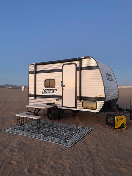 Experience beach camping with this Coleman rv rental. Setup includes a generator, picnic table, and rug on sandy ground under a clear sky.