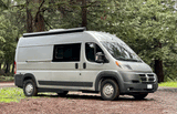 This spacious light gray RV rental camper van is parked on a forest trail, ready for your next nature road trip.