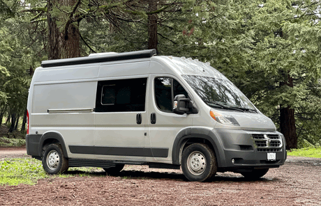 This spacious light gray RV rental camper van is parked on a forest trail, ready for your next nature road trip.