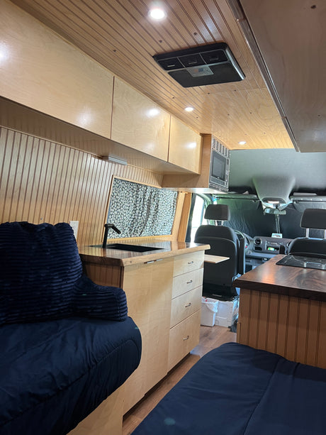 Comfortable camper van rv rental interior with light wood, kitchenette, overhead AC, and dark blue beds.