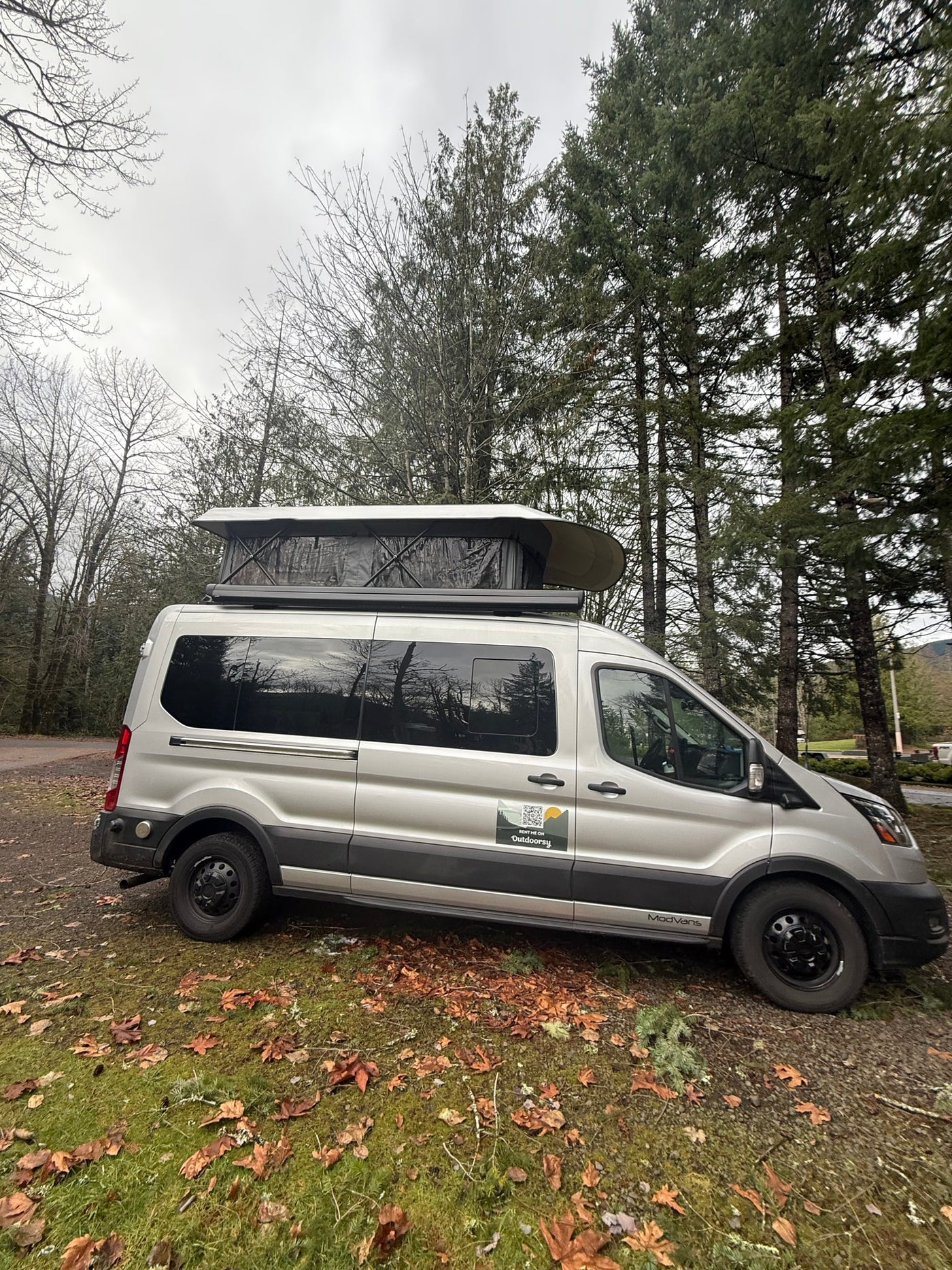 This silver Ford Transit RV rental, with a pop-top camper, is parked in a wooded setting, ready for outdoor travel. — photo 13