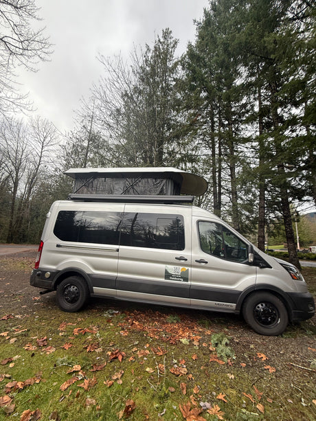 This silver Ford Transit RV rental, with a pop-top camper, is parked in a wooded setting, ready for outdoor travel.