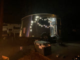 Nighttime RV rental scene: An Airstream-style camper lit with string lights, a person inside, and a dog outside.