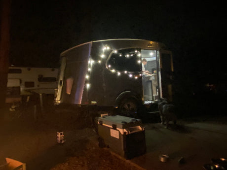 Nighttime RV rental scene: An Airstream-style camper lit with string lights, a person inside, and a dog outside.