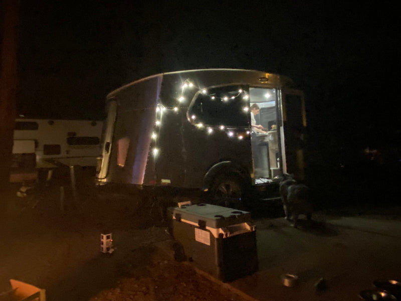 Nighttime RV rental scene: An Airstream-style camper lit with string lights, a person inside, and a dog outside. — photo 7