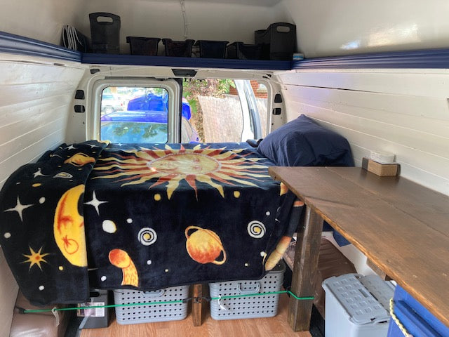 Cozy camper van interior, ideal for an RV rental. Features a large bed with a unique space-themed blanket, storage, and a handy table.