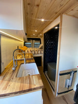 Explore this stylish rv rental camper van's interior: a wooden kitchen, cozy bed, and compact tiled bathroom.