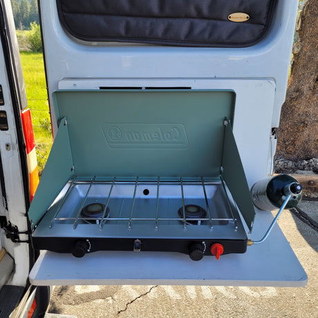 RV rental van featuring a pull-out table and Coleman camp stove with propane tank, ideal for outdoor cooking.