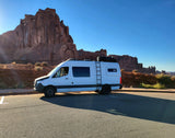 An rv rental, a white camper van, is parked against a backdrop of towering red rock formations in a sunny national park.