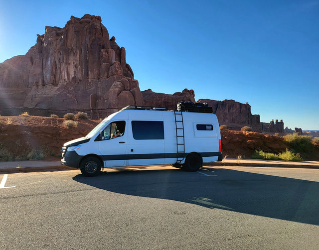 An rv rental, a white camper van, is parked against a backdrop of towering red rock formations in a sunny national park.