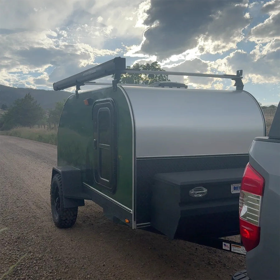 This green teardrop camper, perfect for RV rental adventures, is ready for off-road travel on a dirt road under a cloudy sky.