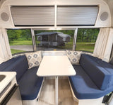 Inside a bright rv rental, a dining area with blue bench seats, a white table, and a scenic view of a campsite and another camper.