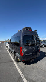 Dark gray Mercedes Sprinter Tranquility camper van RV rental with a bike rack, parked in a sunny outdoor lot.