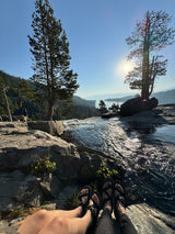 Feet up, relaxing by sparkling water flowing over rocks, with a sunny lake & mountains in the distance.