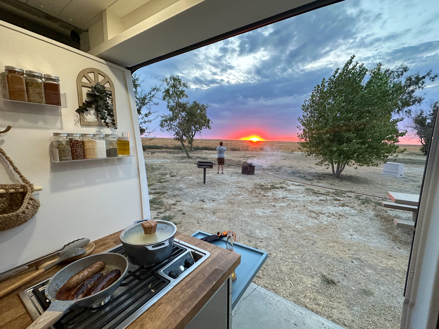 RV rental sunset cooking: sausages sizzle while watching vibrant outdoor views from the camper kitchen.