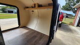 Interior of an rv rental, featuring wood-look flooring, a shelf, and open door view. Outside, a red truck and building are visible.