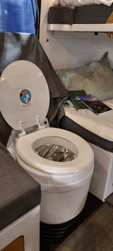 **RV rental** interior: a white composting toilet with its lid open, revealing a foil-lined bowl. A warning sticker is on the lid.