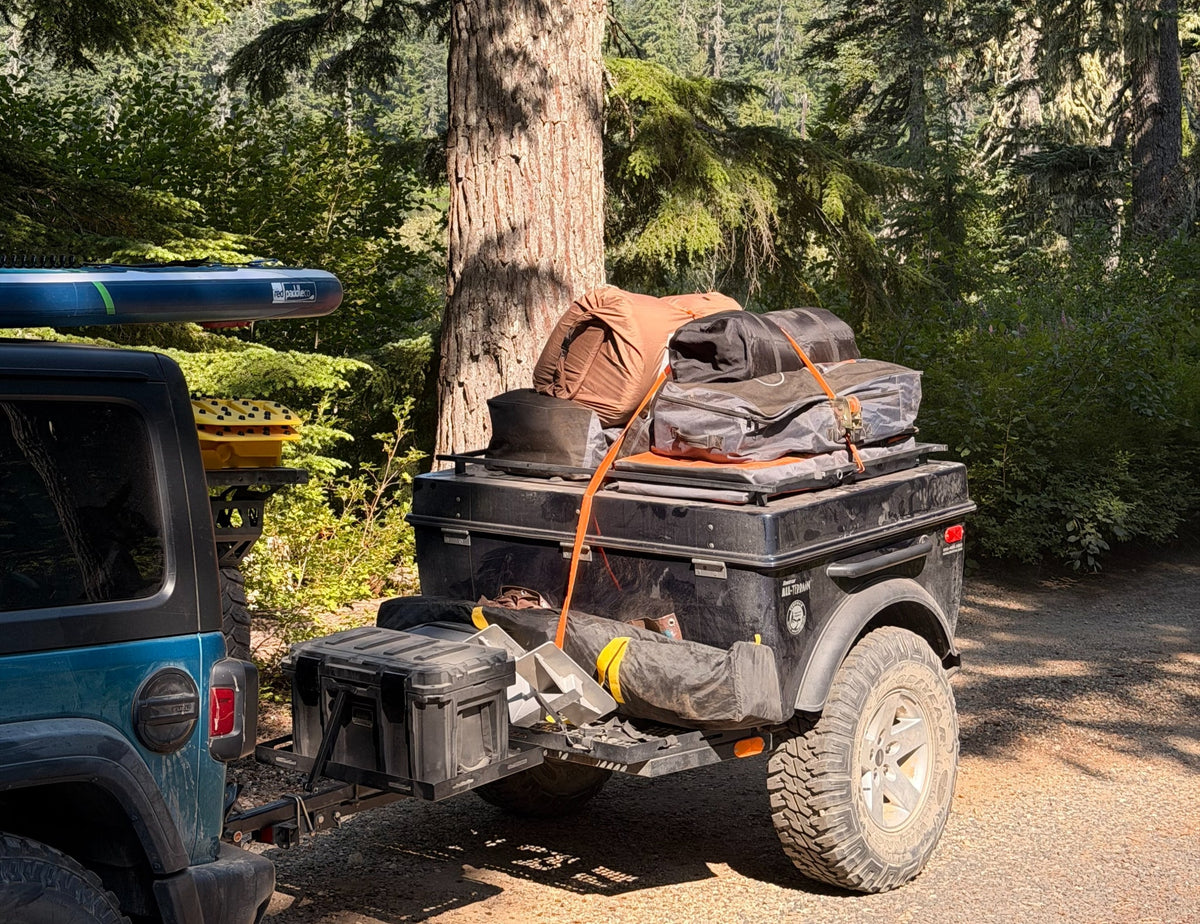 This off-road camping setup, featuring a Jeep, trailer, paddleboard & gear, offers outdoor adventure like an rv rental.