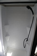 RV rental's bright, compact shower interior with white faux subway tile walls, corner shelves, and a handheld shower.