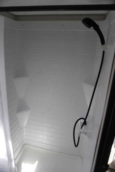 RV rental's bright, compact shower interior with white faux subway tile walls, corner shelves, and a handheld shower.