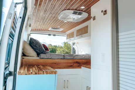 Inside a bright RV rental: view of the comfy bed, wooden ceiling, and compact kitchen/living area with AC unit.