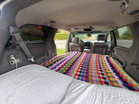 View from rear of a minivan converted for car camping or an RV rental. A comfortable bed with white bedding and a colorful crochet blanket fills the back.