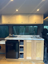 Compact rv rental kitchen: light wood cabinets, counter, sink, built-in fridge. Rear windows show outdoor forest scene.
