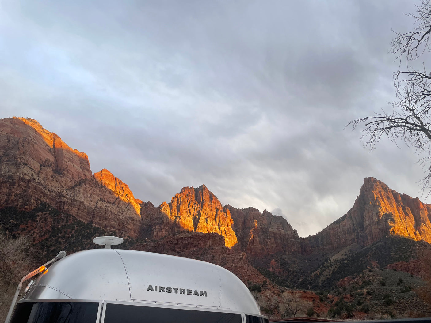 Silver Airstream RV rental parked before sunlit red rock mountains under a cloudy sky. Explore national parks by RV!