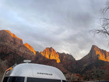 Silver Airstream RV rental parked before sunlit red rock mountains under a cloudy sky. Explore national parks by RV!