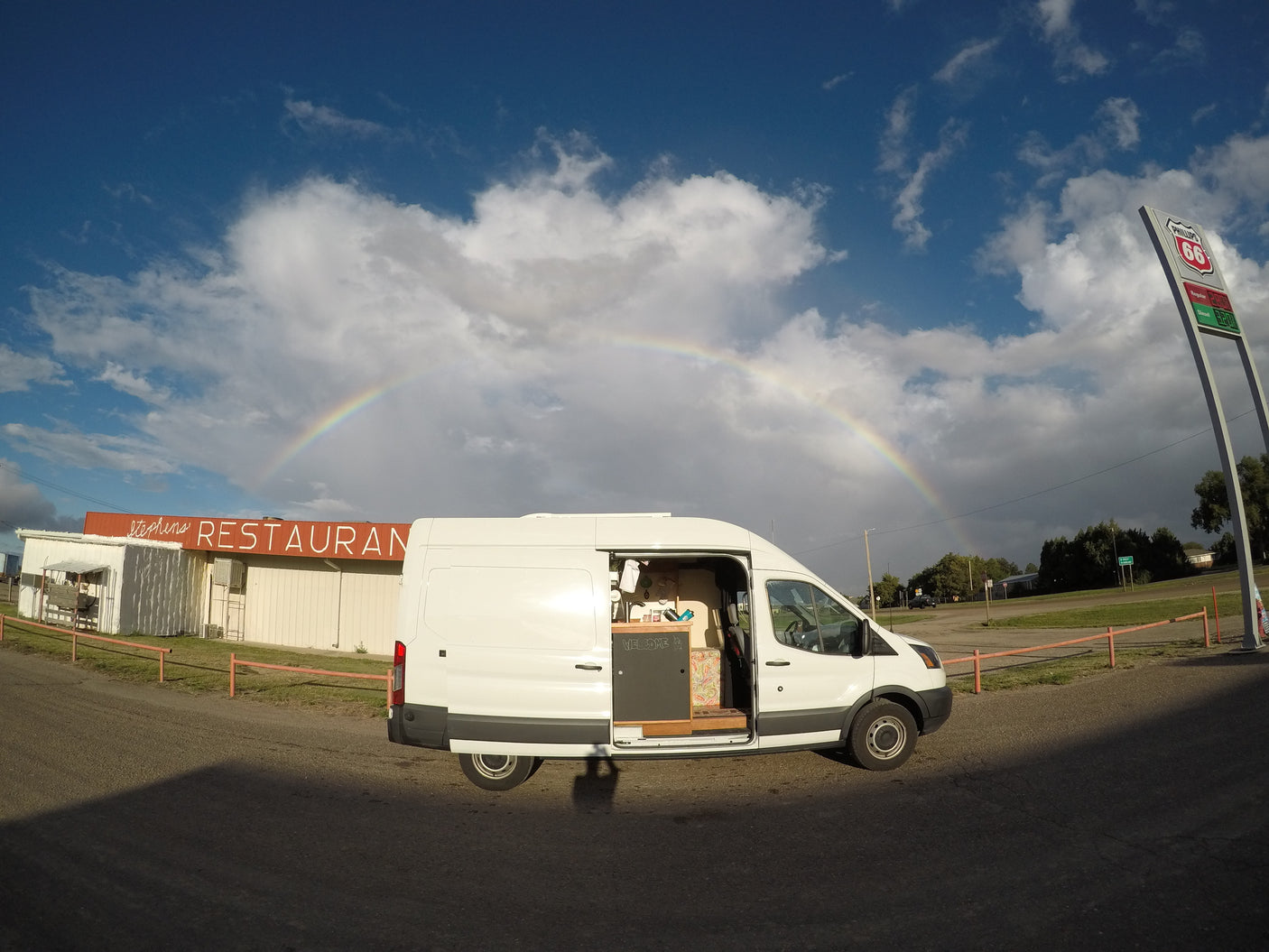 White camper van (RV rental) under a vibrant rainbow near Stephens Restaurant and Phillips 66 sign.
