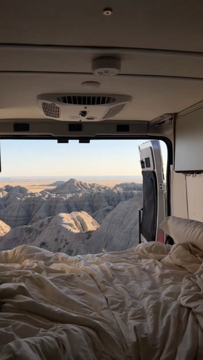 Cozy RV rental bed with open doors revealing epic badlands. Interior shows AC unit, smoke detector. Perfect for scenic travel!