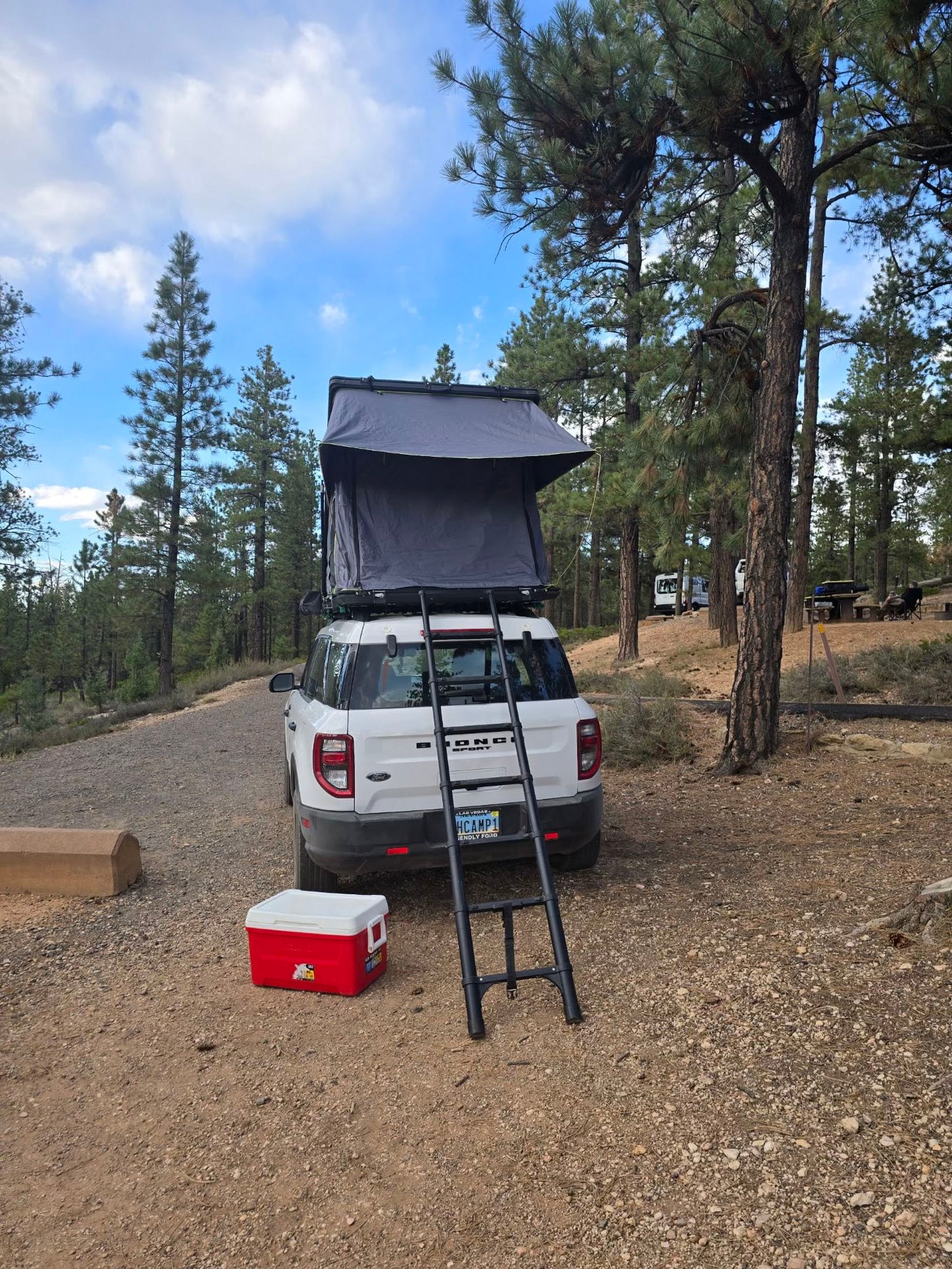 RV rental: A white Bronco Sport with a rooftop tent & ladder, parked at a forested campsite with a cooler.
