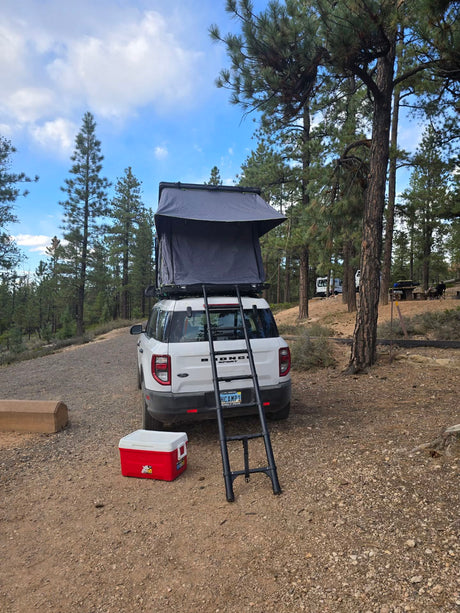 RV rental: A white Bronco Sport with a rooftop tent & ladder, parked at a forested campsite with a cooler.