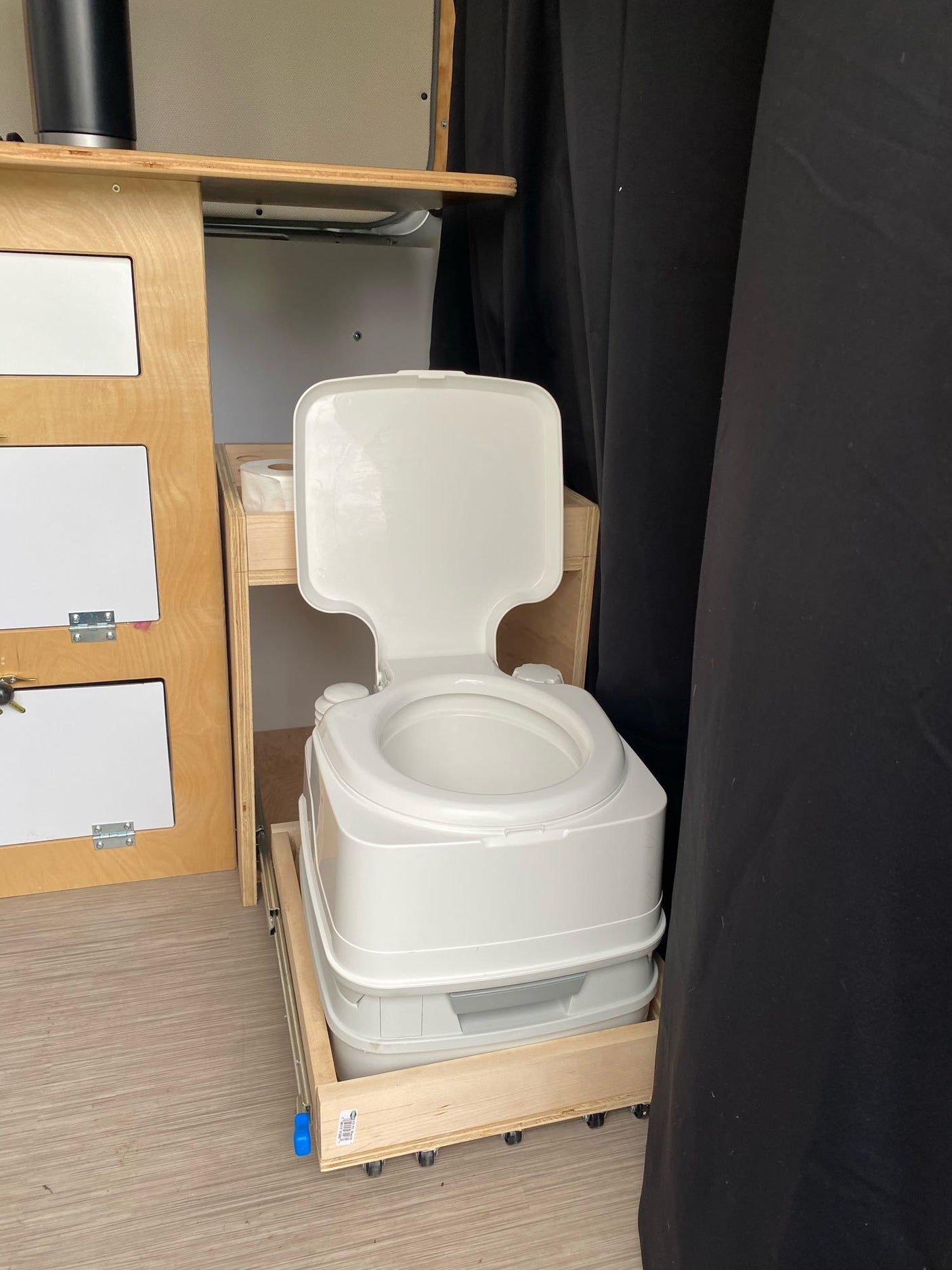 RV rental essential: A white portable toilet on a pull-out base is positioned in a camper's compact interior, by wooden storage.