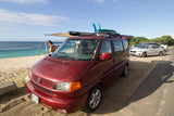 Red VW camper van with surfboards at a Hawaii beach, awning out. Woman stands by. Ideal RV rental for ocean fun.