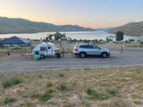 RV rental: Silver SUV towing a teardrop trailer set up at a scenic lakeside campsite with mountains under a soft sky.