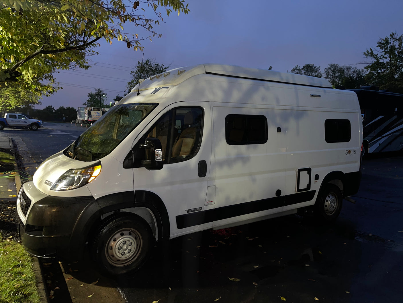 White Winnebago Solis camper van for RV rental, parked on a wet lot at dusk, ready for adventure.