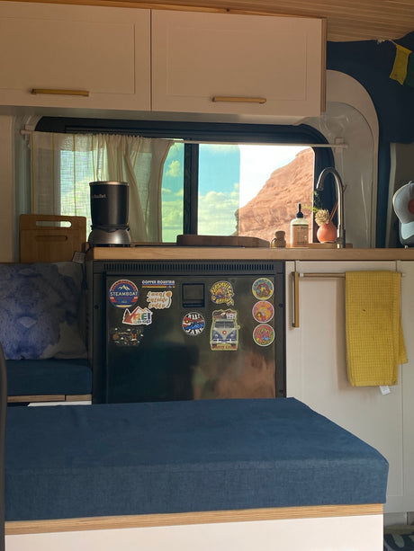 Inside an RV rental: a compact kitchen with a sticker-covered fridge, sink, and desert view. Blue cushions invite relaxation.