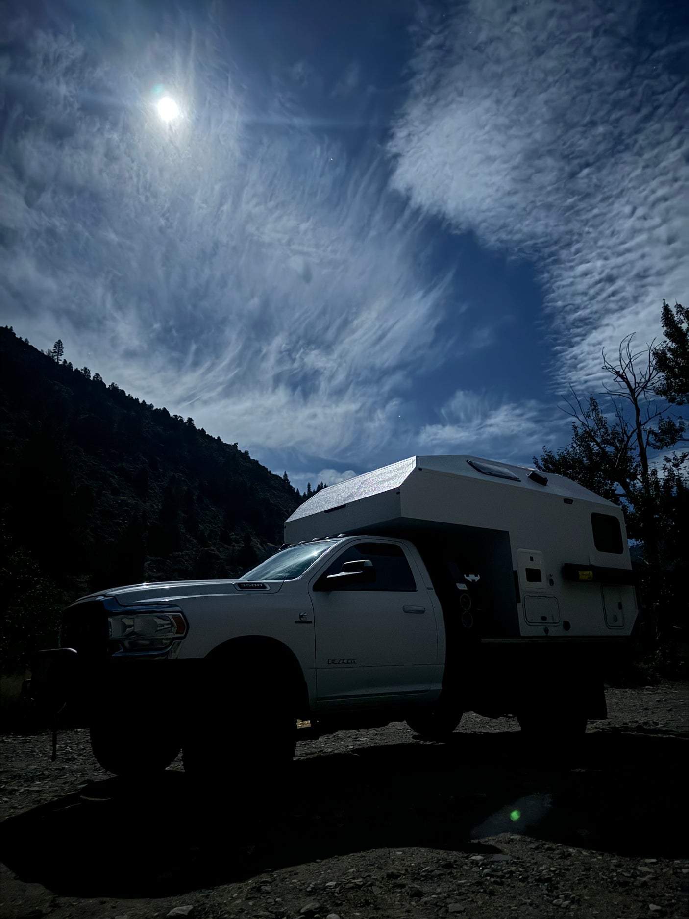 An overlanding truck camper RV rental parked beneath a bright sun and swirling clouds in a dark mountain landscape.