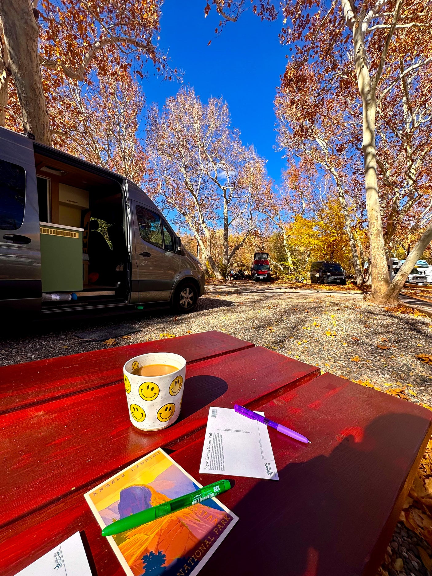 Our RV rental parked among vibrant fall trees at a national park campsite. Coffee & postcards on the bright red picnic table.