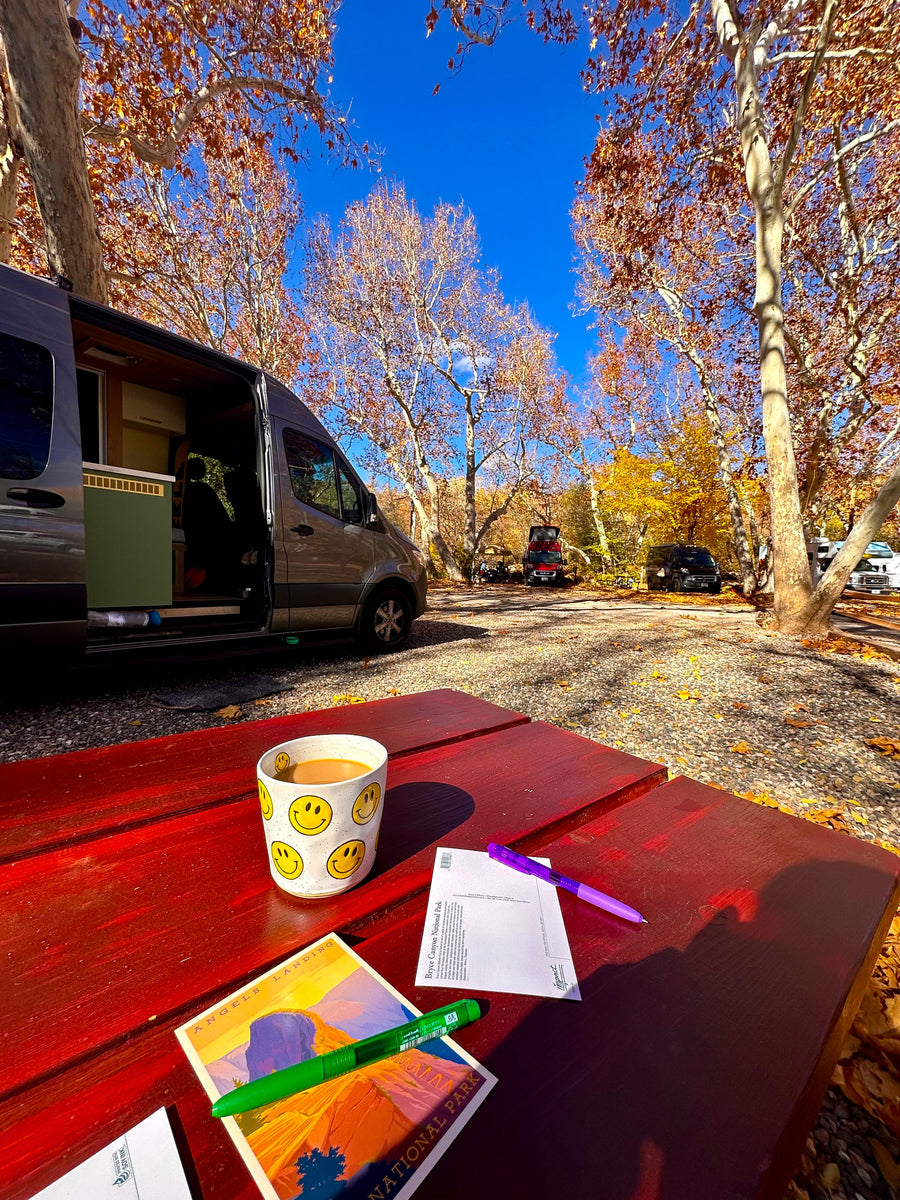 Our RV rental parked among vibrant fall trees at a national park campsite. Coffee & postcards on the bright red picnic table.