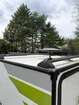 RV rental's roof with satellite dish & racks, green-striped side visible. Pine trees in background under cloudy sky.