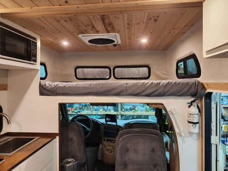 This RV rental features a cozy wooden ceiling, upper bunk bed, kitchenette, and view into the driver's cab. A fire extinguisher is visible for safety.