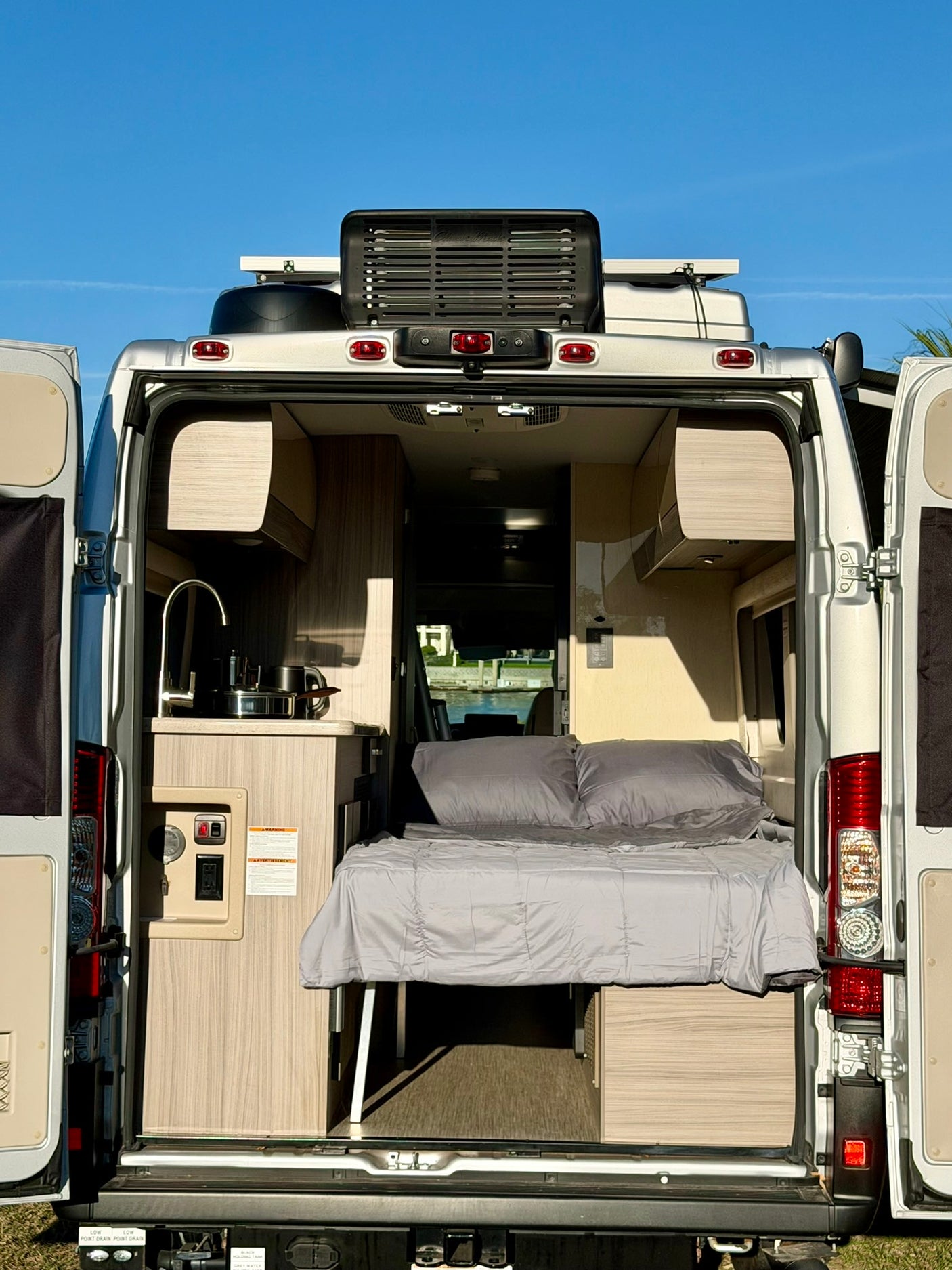 Open rear doors of this RV rental camper van show a compact interior: kitchen with sink, and a neatly made bed, ready for your trip.